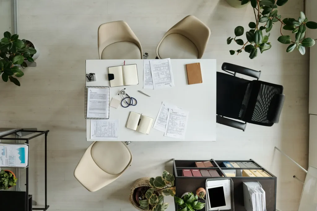 A top down viewpoint of a desk with clipboards, notebooks and stethoscope, surrounded by empty chairs.